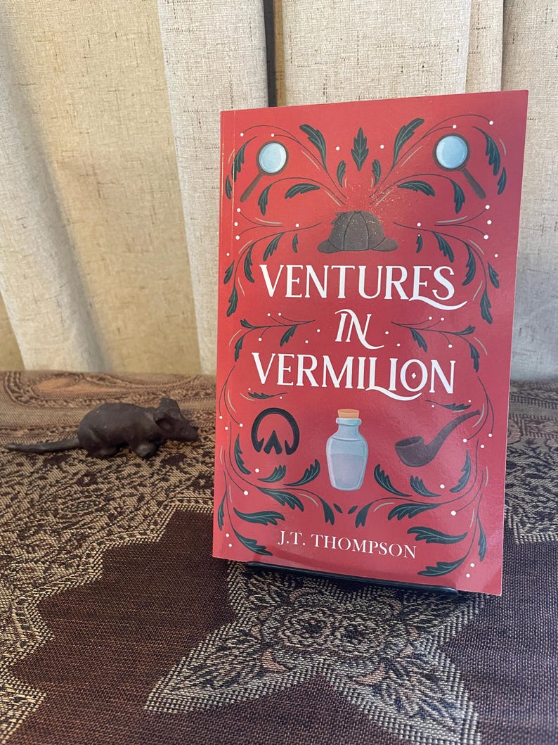 Op de afbeelding: Een boek getiteld "Ventures in Vermilion" met een rode kaft en decoratieve illustraties. De kaft bevat een vergrootglas, een hoed en andere detective-thema-elementen. De naam van de auteur, J.T. Thompson, staat onderaan gedrukt.