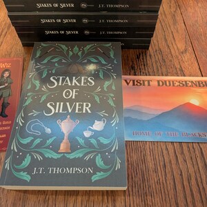 Op de afbeelding: Een stapel boeken Stakes of Silver van J.T. Thompson, een bladwijzer met een personage genaamd Wiz en een ansichtkaart met de tekst Visit Duesenburg Home of the Blackstone Cup. De boekomslag toont een trofee, theepot en ketting.