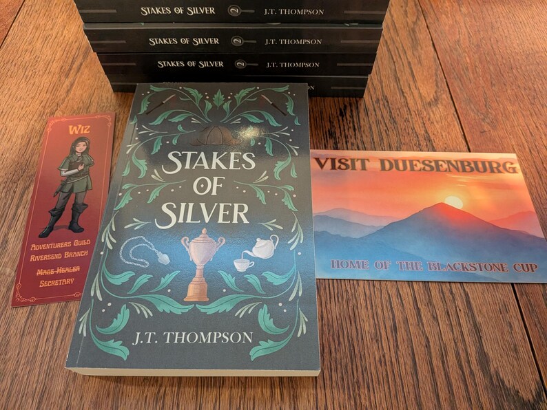 Op de afbeelding: Een stapel boeken Stakes of Silver van J.T. Thompson, een bladwijzer met een personage genaamd Wiz en een ansichtkaart met de tekst Visit Duesenburg Home of the Blackstone Cup. De boekomslag toont een trofee, theepot en ketting.