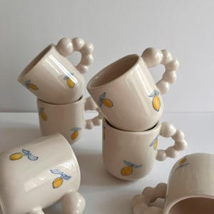 May include: Set of five white ceramic mugs with a lemon design. The mugs have a unique beaded handle.