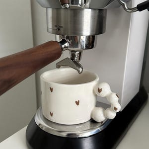 Può includere: Una macchina da caffè espresso bianca con un portafiltro argentato e un manico in legno marrone. Una tazza in ceramica bianca con disegni di cuori marroni e un'unica maniglia perlata è posizionata sul vassoio di gocciolamento.