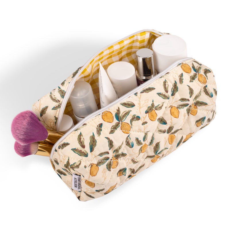 May include: A white quilted makeup bag with a lemon print and a yellow and white checkered lining. The bag is open and filled with various beauty products, including bottles, tubes, and brushes.