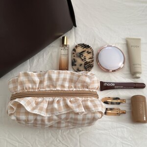 Può includere: Una trousse per il trucco a quadri beige e bianchi con un bordo a balze e una cerniera dorata. La trousse è circondata da vari prodotti di bellezza, tra cui una bottiglia di olio per il viso Nuxe, uno specchio compatto, un tubo di crema idratante PUSY, un tubo di trattamento labbra rhode e due fermagli per capelli dorati.