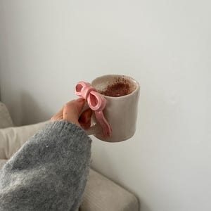 Puede incluir: Una taza de cerámica blanca con un asa en forma de lazo rosa, llena de una bebida marrón claro. La taza está siendo sostenida por una mano que lleva un suéter gris.