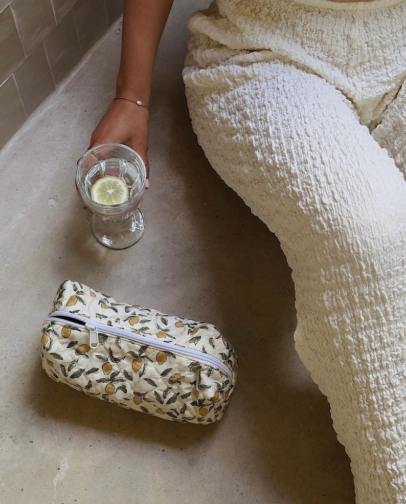 May include: A white zippered pouch with a lemon print design. The pouch is lying on a light brown surface.