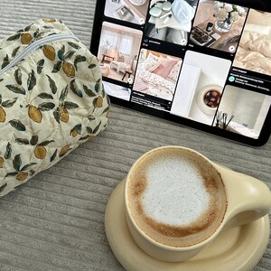 May include: A close-up of a cup of cappuccino with a light brown ceramic mug and saucer. The mug has a rounded handle and the saucer is slightly larger than the mug. The cappuccino has a thick layer of foam on top. The cup is sitting on a gray textured surface.
