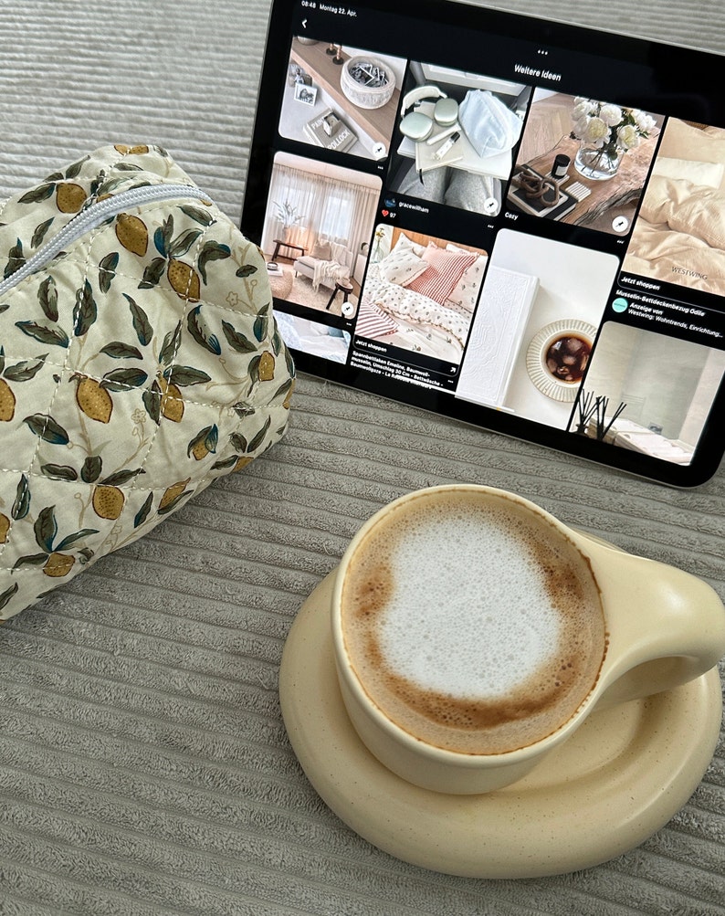 May include: A close-up of a cup of cappuccino with a light brown ceramic mug and saucer. The mug has a rounded handle and the saucer is slightly larger than the mug. The cappuccino has a thick layer of foam on top. The cup is sitting on a gray textured surface.