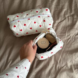 May include: Two white quilted cosmetic bags adorned with red hearts. One bag is open, displaying makeup items. The bags feature white zips and are placed on a neutral surface. One bag is being held open.