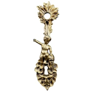 Op de afbeelding: Oorspronkelijk goudkleurig deurbeslag met een cherubijnfiguur. De cherubijn houdt een vat vast, met een sleutelgat eronder. Het beslag heeft een decoratief, bloemig ontwerp.