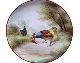 May include: A round ceramic plate with a red rim features a painted scene of two roosters in a green and brown landscape. The rooster in the foreground is colorful with red, yellow, blue, and green feathers. The rooster in the background is black.