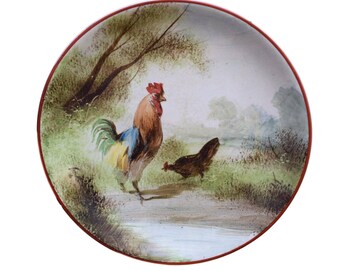 May include: A ceramic plate with a hand-painted design of a rooster and a hen in a green and brown landscape. The plate has a red rim.