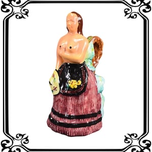 Könnte beinhalten: Keramikfigur einer Frau mit teilweise nacktem Oberkörper, die einen rot gestreiften Rock mit schwarzem Besatz und eine schwarze Schürze mit gelben floralen Details trägt. Die Figur hat braunes Haar und einen Korb auf dem Rücken.