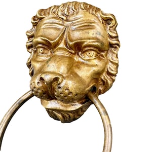 May include: A gold-coloured lion head door knocker with a ring handle. The lion's face is detailed with a furrowed brow and textured mane. The ring handle is attached to the lion's mouth.