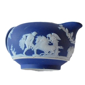 May include: A blue ceramic creamer with white bas-relief figures and floral designs. The creamer features a handle and a spout. The bas-relief depicts classical figures and a chariot. The creamer is a decorative piece.