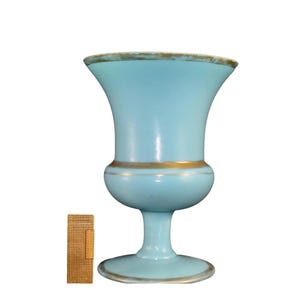 May include: A light blue ceramic vase with a gold rim and a gold band around the middle. The vase has a classic urn shape with a wide base and a narrow neck.