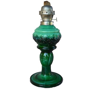 Könnte beinhalten: Eine grüne Glas-Öllampe mit einem verzierten Design und einem Metall-Docht-Halter. Die Lampe hat einen runden Sockel und einen hohen, schlanken Körper.