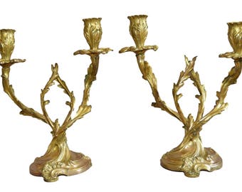 May include: A pair of ornate gold-toned metal candle holders with a floral design. Each holder has three candle cups and a decorative base.