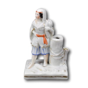 May include: A white porcelain figurine depicting a person in a white coat, blue and white striped shorts, and a red sash. The figurine stands beside a white cylindrical structure. The figurine is on a rectangular base.