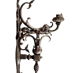 Puede incluir: Una pared de metal marrón oscuro con un diseño floral y un solo portavelas. La pared tiene un diseño decorativo de volutas y una cabeza de pájaro en la parte superior.