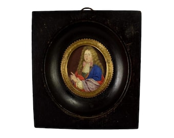 May include: A framed miniature portrait of a man with long, curly hair, wearing a blue coat and a pink cravat. The portrait is set within a gold-colored, circular frame, encased in a dark, square wooden frame. The artwork is set against a dark red background.