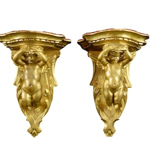 May include: A pair of ornate gold-painted wall sconces, each featuring a cherub sculpture with wings, holding its hands over its eyes. The sconces have a decorative, scrolled design.