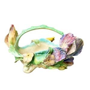 May include: A ceramic basket-shaped planter with a handle, adorned with sculpted flowers and leaves. The planter features pastel colors including green, yellow, blue, and purple. The floral design is detailed and adds a decorative touch.