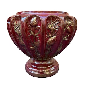 Puede incluir: Un macetero de cerámica rojo con un diseño pintado en oro de piñas y ramas. El macetero tiene una base ancha y redonda y un cuello estrecho y cilíndrico.