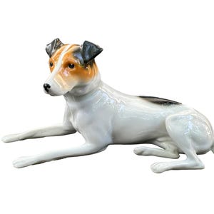 May include: A ceramic dog figurine in a relaxed pose. The dog has a white body with brown and black markings on its head and back. The figurine is glossy and appears to be a decorative item.