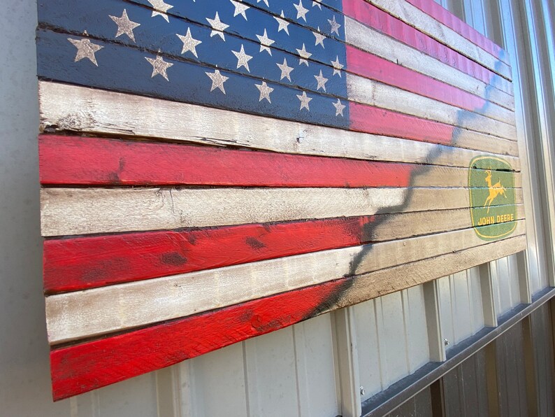 May include: Wooden American flag with red and white stripes and a blue field with white stars. The flag has a distressed look and features the John Deere logo in the lower right corner.