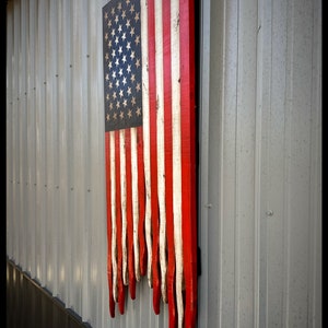 May include: A wooden American flag with red, white and blue stripes. The flag is hanging on a wall and has a distressed look. The stripes are painted on individual pieces of wood and are dripping down the wall.