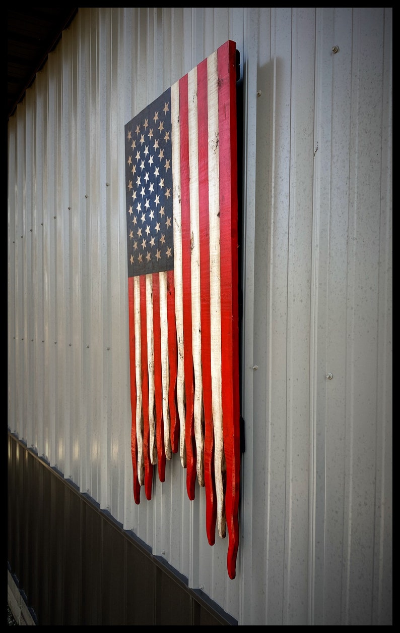 May include: A wooden American flag with red, white and blue stripes. The flag is hanging on a wall and has a distressed look. The stripes are painted on individual pieces of wood and are dripping down the wall.