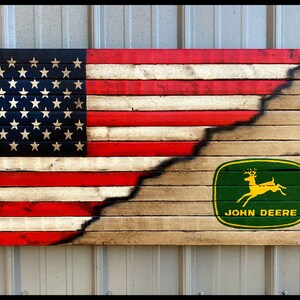 May include: Wooden wall art depicting the American flag design, with the stars and stripes on the left and a John Deere logo on the right. The flag is painted on wooden planks, with a distressed finish. The dimensions are approximately 76 cm wide and 38 cm tall.