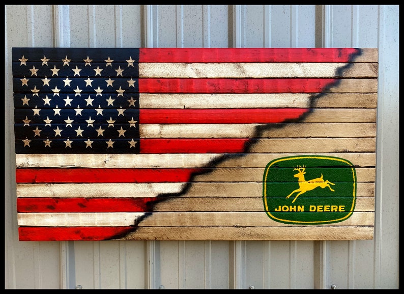 May include: Wooden wall art depicting the American flag design, with the stars and stripes on the left and a John Deere logo on the right. The flag is painted on wooden planks, with a distressed finish. The dimensions are approximately 76 cm wide and 38 cm tall.