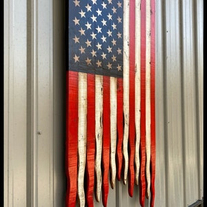 May include: A wooden American flag painted in red, white and blue. The flag is hanging on a wall and the bottom of the flag is frayed and uneven.