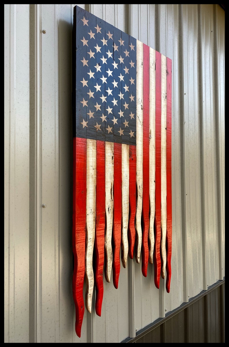 May include: A wooden American flag painted in red, white and blue. The flag is hanging on a wall and the bottom of the flag is frayed and uneven.