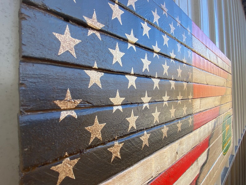 May include: Wooden wall art depicting the American flag. The design includes gold stars on a black field, red and white stripes, and a red line. The artwork is constructed from wooden planks, offering a rustic aesthetic.