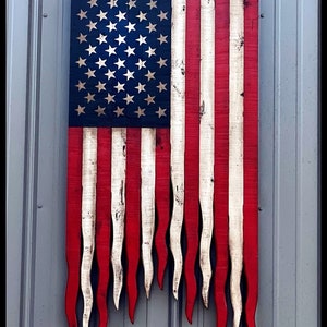 May include: A wooden American flag with a distressed finish. The red and white stripes are frayed at the bottom, giving the flag a tattered look. The blue field with white stars is intact.