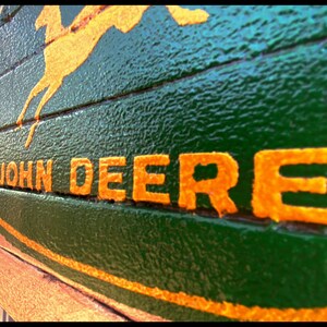 May include: A green wooden surface with the John Deere logo painted on it. The logo includes a leaping deer and the words "JOHN DEERE" in orange. The background is a textured green.