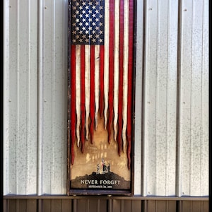 May include: A wooden American flag with red, white, and blue stripes. The flag is distressed and has a weathered look. The flag is hanging on a wall with a metal siding. The flag has a scene of firefighters at the World Trade Center with the text "NEVER FORGET SEPTEMBER 11, 2001".