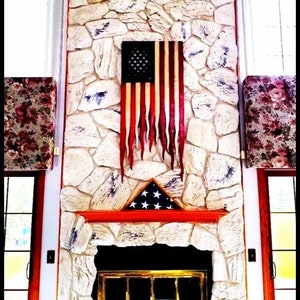 May include: A weathered American flag hangs above a fireplace with a stone surround. The flag is tattered and has a distressed look. The fireplace has a red mantel and a black metal grate.
