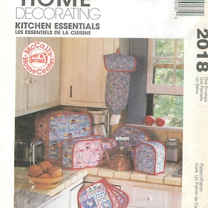 May include: A collection of kitchen essentials, including covers for appliances and pot holders. The covers feature various patterns with red trim. The image also shows the text "McCall's Home Decorating Kitchen Essentials".