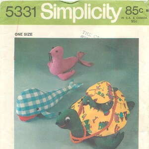 May include: Simplicity pattern 5331 featuring stuffed animal designs. Includes a pink seal, a blue and white checkered whale, and a green turtle with a yellow shell. A child is shown with the stuffed animals.