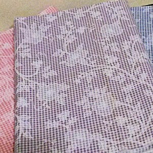 May include: Three folded pieces of fabric featuring a small gingham check pattern in pink, purple, and blue. Each piece of fabric is overlaid with a white, intricate floral vine print that spans across the surface of the checkered material.