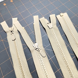 May include: Five beige zippers of varying lengths laid out on a gray gridded surface. The zippers have a metal slider and teeth.