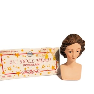 May include: A 2 1/2 inch porcelain doll head with brown hair and a neutral facial expression. The doll head is packaged on a card with the text "Doll Head Porcelain" and "Fibre Craft".