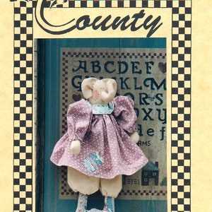 Peut inclure: Une poupée en tissu faite à la main portant une robe rose et un tablier à motifs bleu clair et blanc. La poupée a un nez rose et un col bleu clair. La poupée est debout devant un fond bleu et blanc avec les lettres de l'alphabet imprimées dessus. La poupée est étiquetée "Lollie".