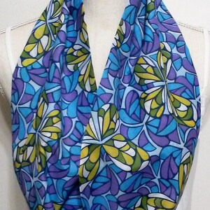 May include: A colorful infinity scarf with a floral pattern. The scarf features a repeating design of stylized leaves in shades of blue, purple, yellow, and green. The scarf is displayed on a mannequin.