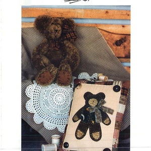 May include: A brown teddy bear sitting on a white crocheted doily. The bear is wearing a red and black plaid bow. A pillow with a brown bear applique is in the background. The pillow is made of patchwork fabric with a cream background. The text "#136 Benjamin Bear" is at the bottom of the image.
