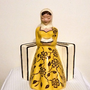 May include: A ceramic figurine of a woman in a yellow dress with a floral pattern, holding a book. The figurine has a white head covering and is positioned in front of two book-shaped supports. The books are white with black trim.