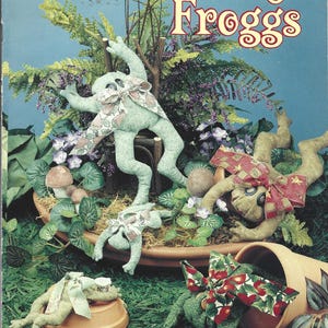 May include: A McCall's pattern cover featuring the title "Friendly Froggs." The image showcases several plush frog figures in a garden setting. The frogs are light green and tan, adorned with patterned bows, and surrounded by greenery and flowers.